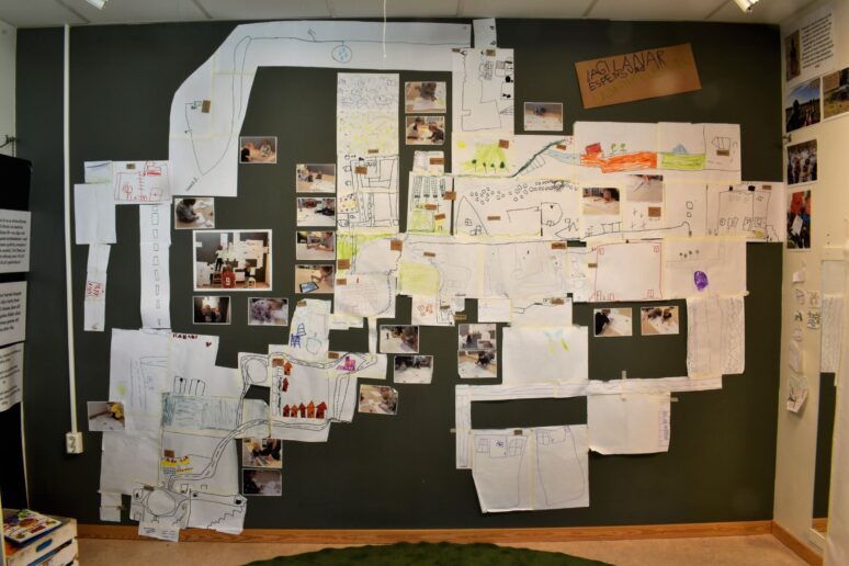 Barns teckningar, kartor och fotografier dokumenterar ett årsprojekt om vägar och platser på en vägg i förskolan.